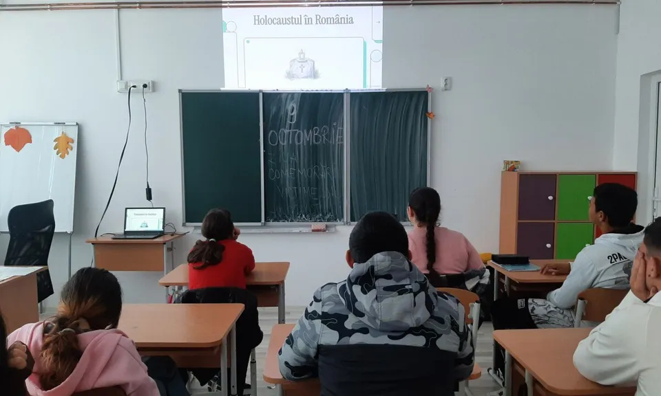 Mai mulți elevi stau la mese orientate spre o tablă, iar o imagine este proiectată pe peretele din față.