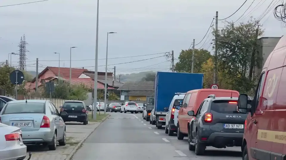 Pe o stradă, mai multe autoturisme și autoutilitare stau în șir; în dreapta se vede o autoutilitară roșie, iar în față, un camion albastru.