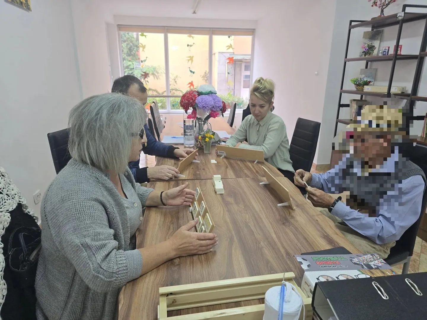 Mai multe persoane stau în jurul unei mese din lemn, cu mâinile lângă materiale mici; pe masă se află flori și cutii.