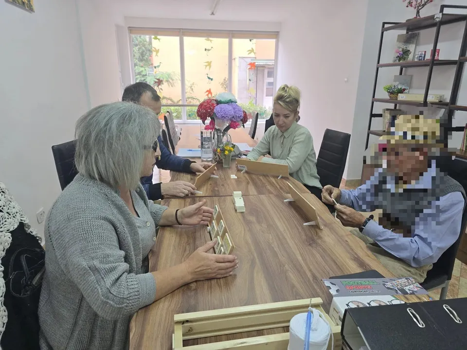 Mai multe persoane stau în jurul unei mese din lemn, cu mâinile lângă materiale mici; pe masă se află flori și cutii.