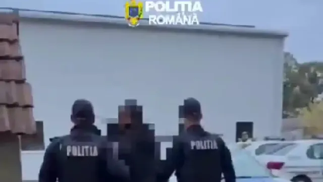 Doi bărbați în uniforme închise la culoare stau de o parte și de alta a unei persoane cu fața pixelată, lângă o clădire și mașini albe.