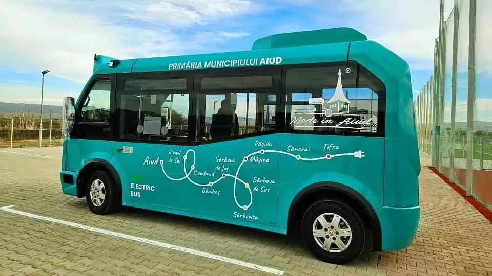 Un autobuz turcoaz, cu geamuri fumurii și marcaje albe, se află pe un pavaj lângă o clădire cu fațadă vitrată.