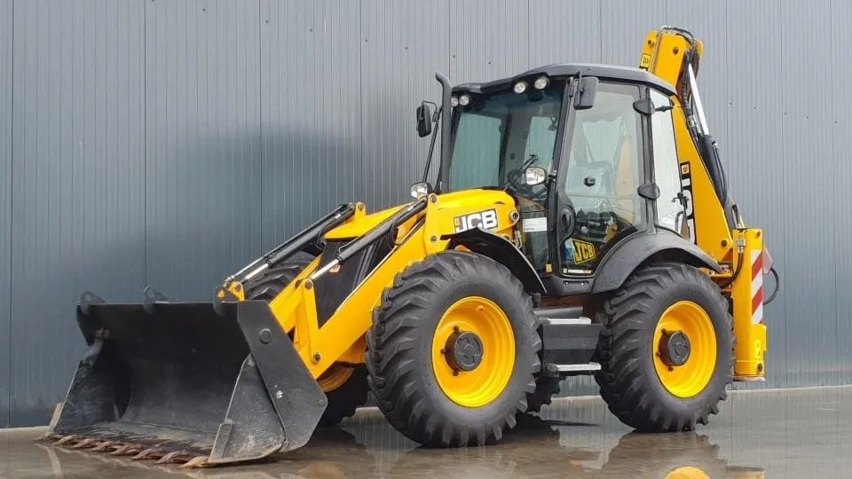 Un buldoexcavator galben cu cabină închisă, cupă frontală și braț posterior este parcat pe o suprafață umedă, lângă un perete gri.