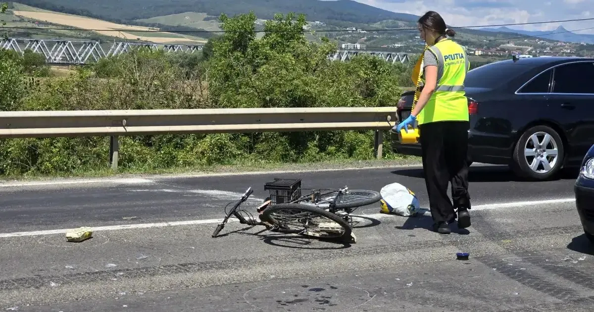 O bicicletă căzută se află pe carosabil, lângă o pungă albă; o persoană cu vestă reflectorizantă stă lângă parapet și o mașină neagră.