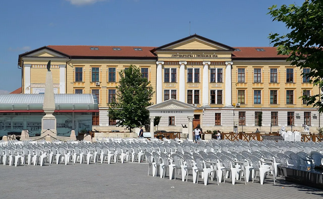În fața unei clădiri galbene cu coloane albe sunt numeroase scaune albe, câteva persoane, un monument înalt și copaci.