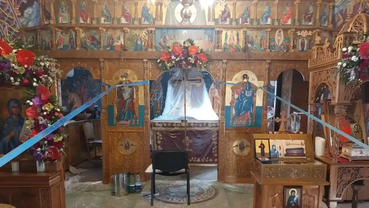 Un iconostas din lemn, cu icoane, este decorat cu flori și panglici albastre; în fața lui se află un scaun negru.
