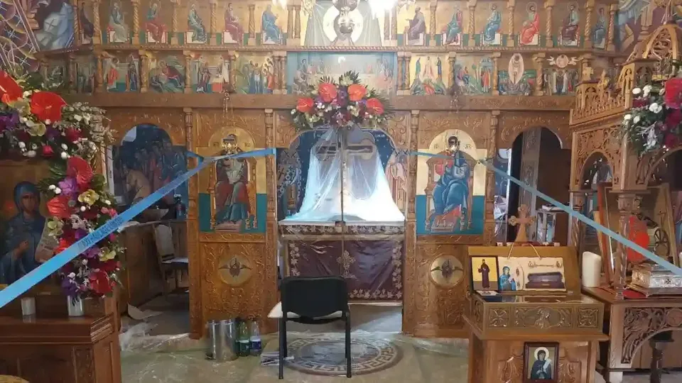 Un iconostas din lemn, cu icoane, este decorat cu flori și panglici albastre; în fața lui se află un scaun negru.