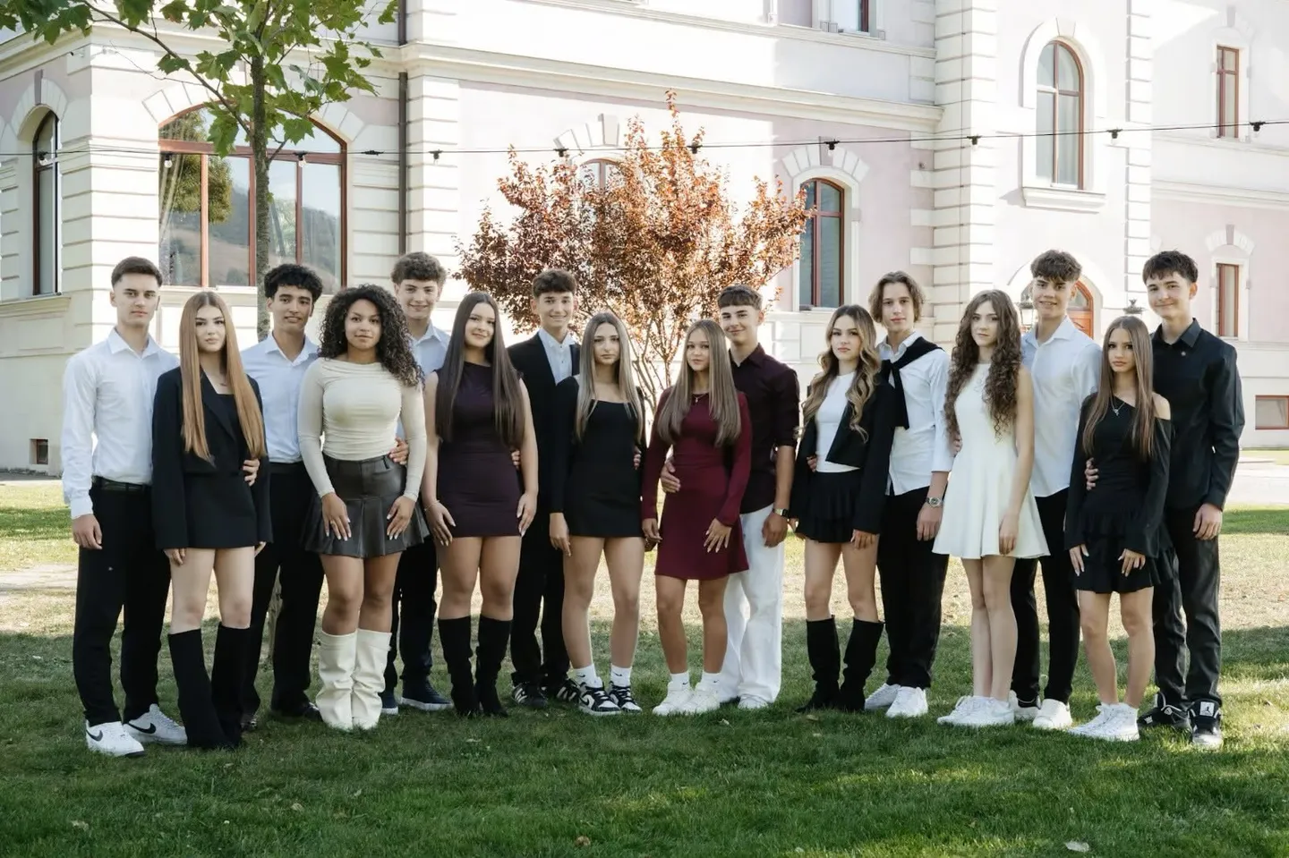 Un grup de tineri stă pe iarbă în fața unei clădiri albe, purtând rochii, cămăși, pantaloni și încălțăminte deschisă la culoare.