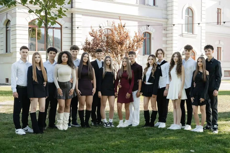 Un grup de tineri stă pe iarbă în fața unei clădiri albe, purtând rochii, cămăși, pantaloni și încălțăminte deschisă la culoare.