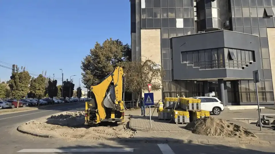 Un utilaj galben stă pe o zonă de pământ excavat, lângă indicatoare, bariere, conuri, un autoturism alb și o clădire modernă.