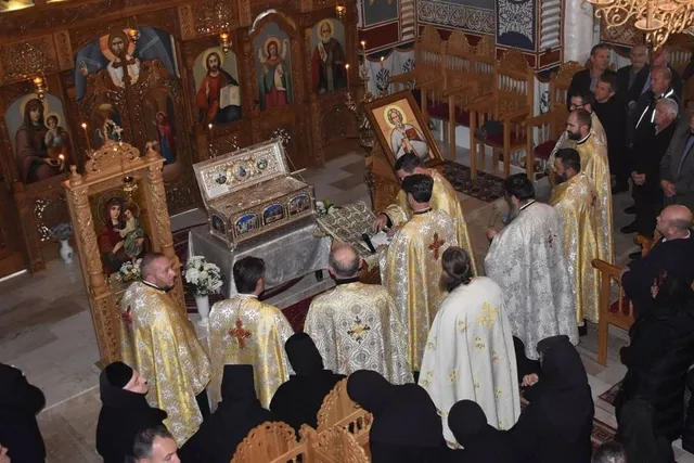 Într-un interior cu icoane, mai multe persoane în veșminte deschise la culoare stau în jurul unei racle ornamentate, alături de alte persoane îmbrăcate în haine închise.