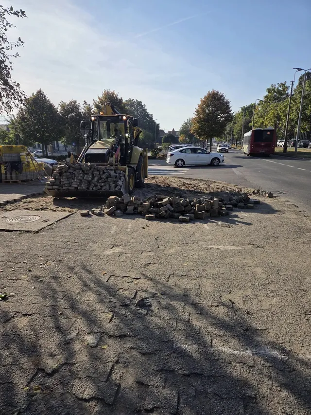 Un utilaj galben stă lângă grămezi de pietre și asfalt spart, iar în fundal se văd o mașină albă, un autobuz și copaci.