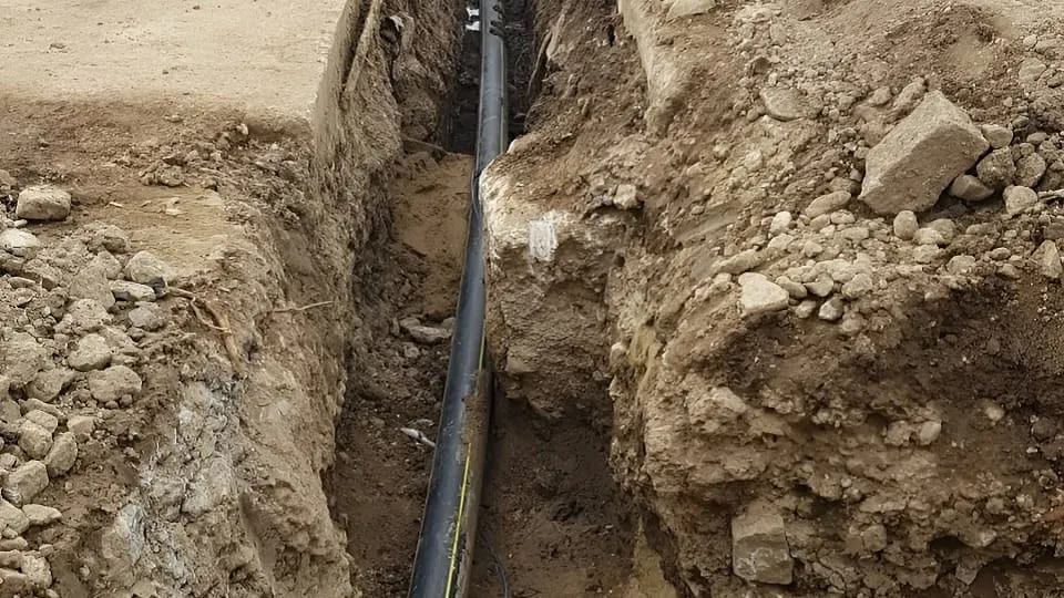 Un șanț îngust traversează solul excavat, iar o conductă neagră se întinde de-a lungul fundului, printre pământ și pietre.