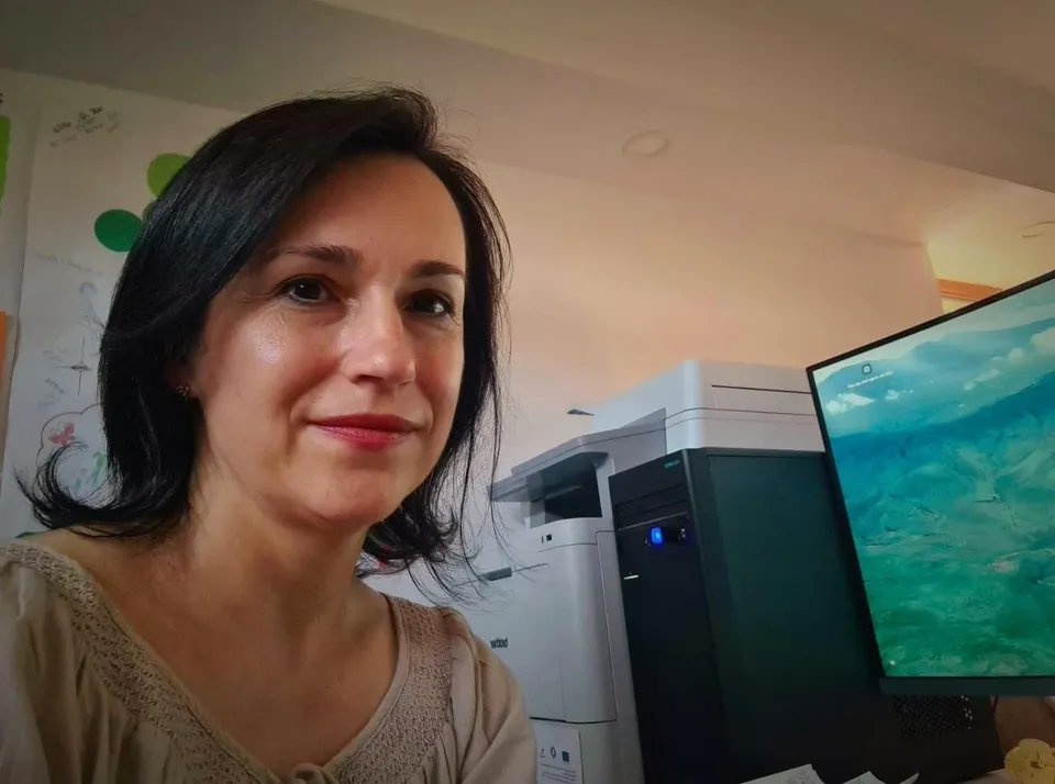 O femeie cu păr închis la culoare și bluză bej se află lângă un monitor și o imprimantă.