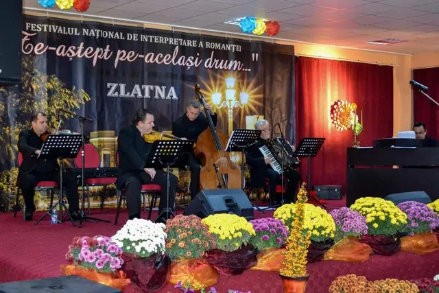 Pe o scenă, mai multe persoane stau lângă pupitre muzicale și instrumente, în fața unei cortine cu text; ghivece cu flori colorate mărginesc scena.