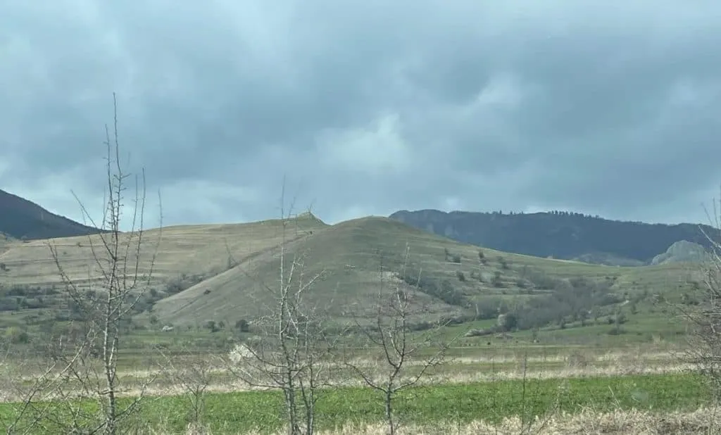 Nori cenușii acoperă cerul deasupra unor dealuri cu vegetație rară; în prim-plan se văd iarbă și copaci fără frunze.
