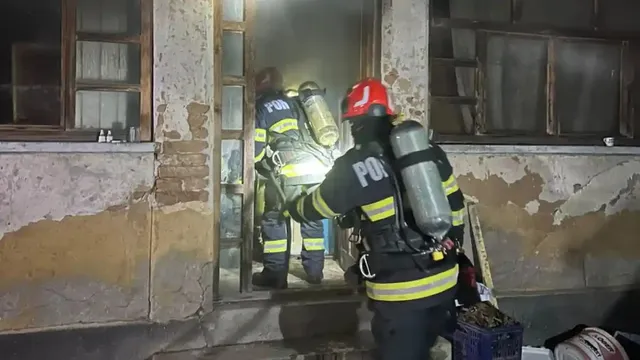 Trei persoane în echipament de protecție și cu butelii pe spate se află lângă intrarea unei clădiri cu pereți deteriorați și ferestre înnegrite.