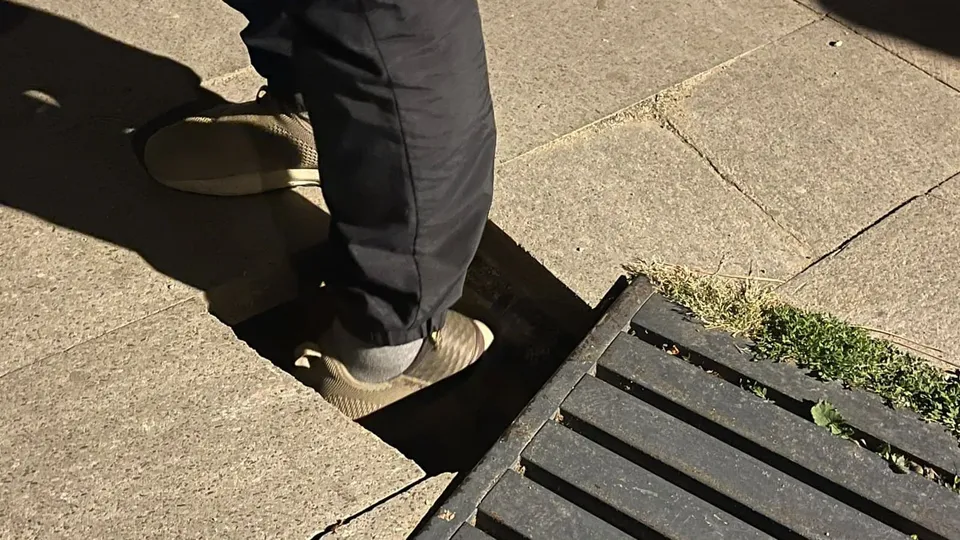 O persoană cu pantaloni negri și pantofi deschiși la culoare stă într-o adâncitură dreptunghiulară, lângă un grătar și dale.