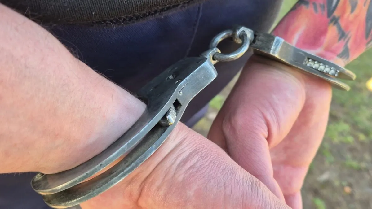 Două mâini sunt apropiate, cu încheieturile prinse în cătușe metalice; pe un antebraț se vede un tatuaj colorat.