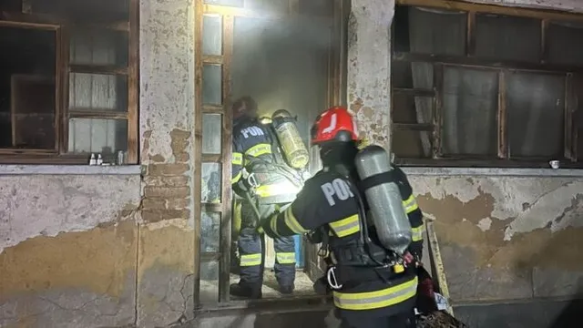 Doi pompieri cu echipament de protecție și butelii pe spate se află lângă intrarea unei clădiri cu pereți deteriorați.