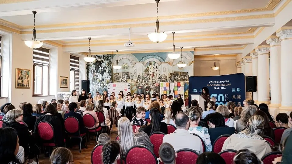Într-o sală decorată, numeroși oameni stau pe scaune orientați spre un grup aflat în picioare, în fața unui panou.