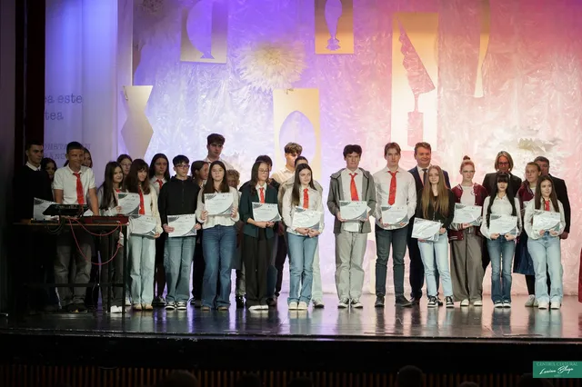 Un grup de tineri și adulți stă pe o scenă, iar mai mulți țin diplome, în fața unui fundal colorat.