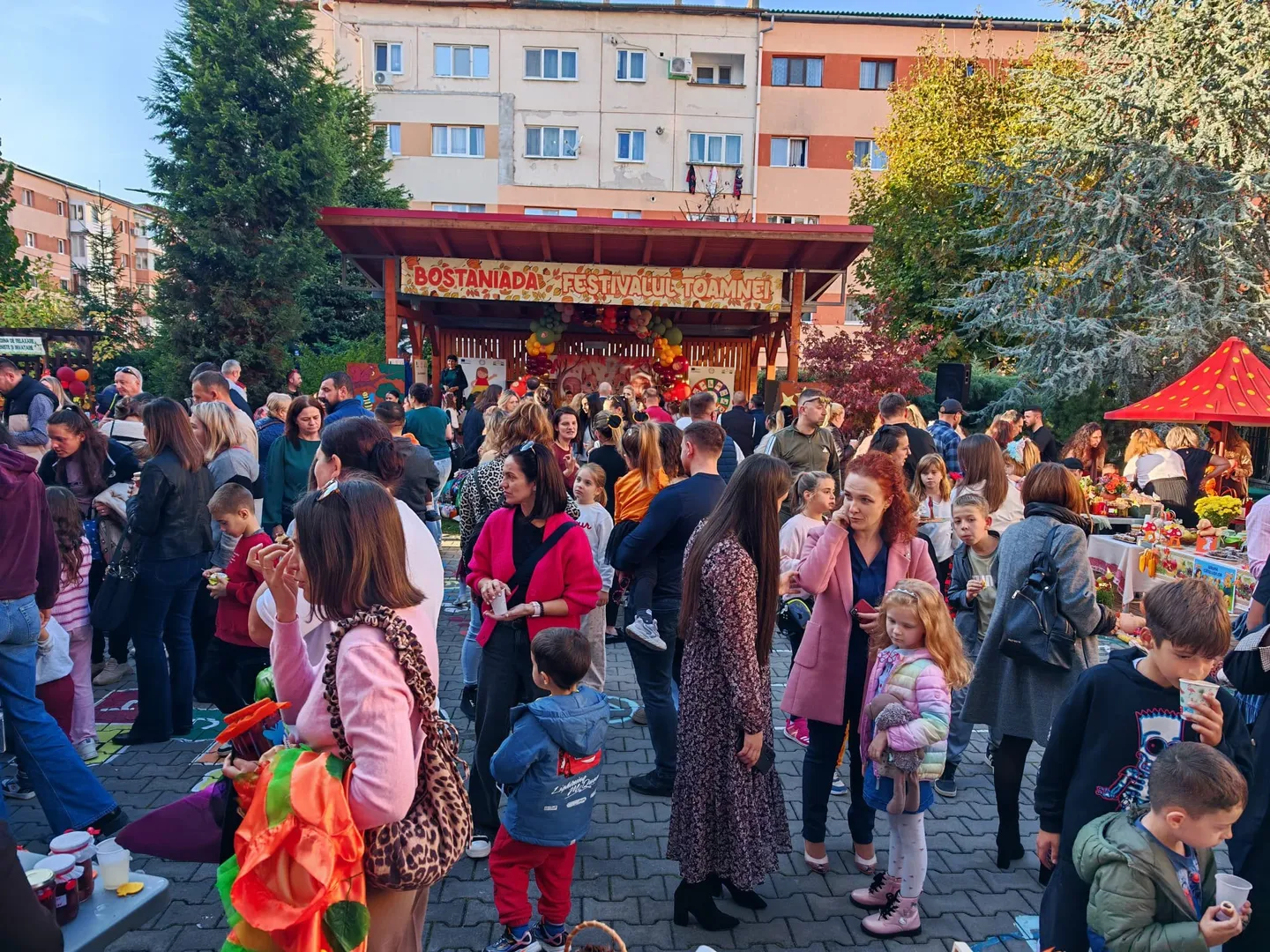 Adulți și copii stau în fața unor standuri decorate cu dovleci și produse colorate, sub un afiș pe care scrie „Bostaniada”.