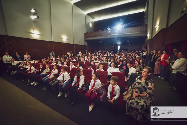 O sală de spectacole este plină de copii în ținute festive, așezați pe scaune, și adulți aflați lateral și în spate.