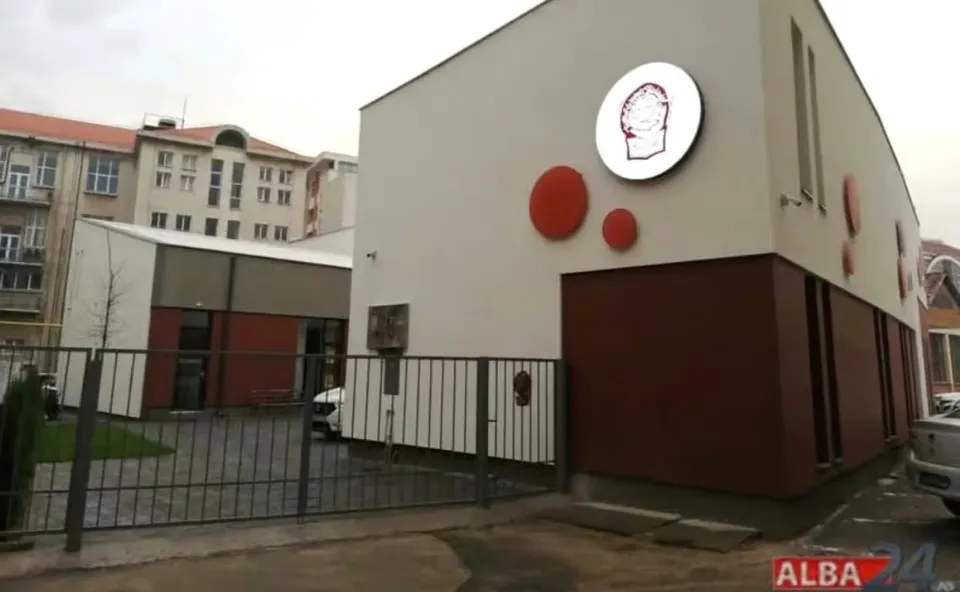 O clădire albă cu panouri maro și cercuri roșii se află în spatele unui gard metalic, lângă blocuri și automobile parcate.