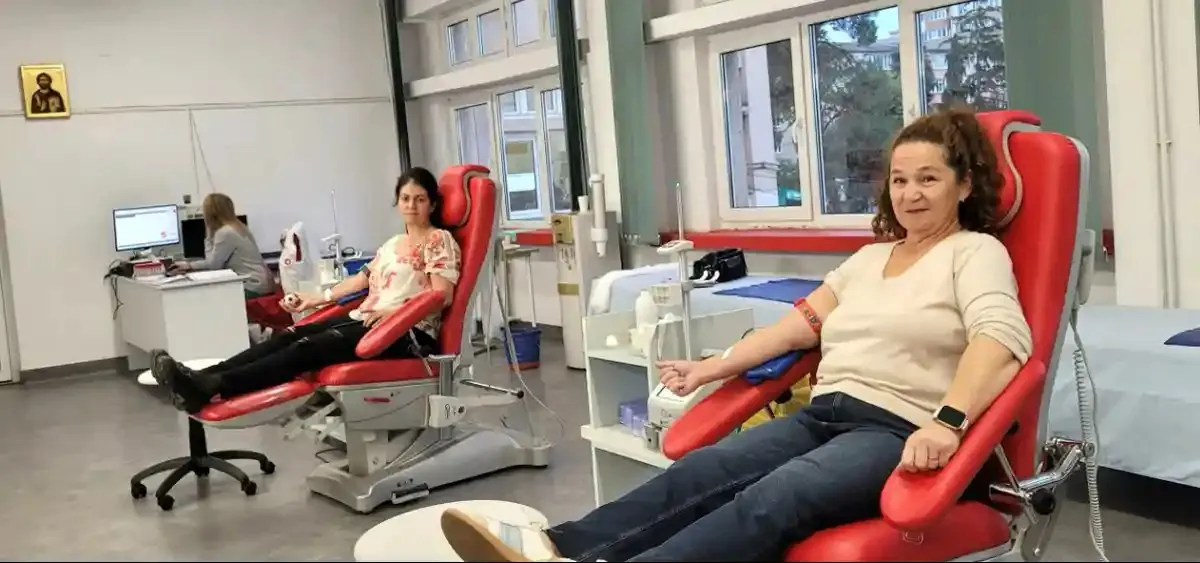 Două femei stau pe scaune roșii înclinate, cu brațele sprijinite pe suporturi; în fundal, o persoană stă la un birou.