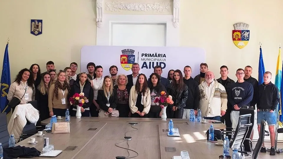 Un grup de persoane stă în spatele unei mese lungi, lângă două buchete de flori, sticle, microfoane, drapele și un panou.