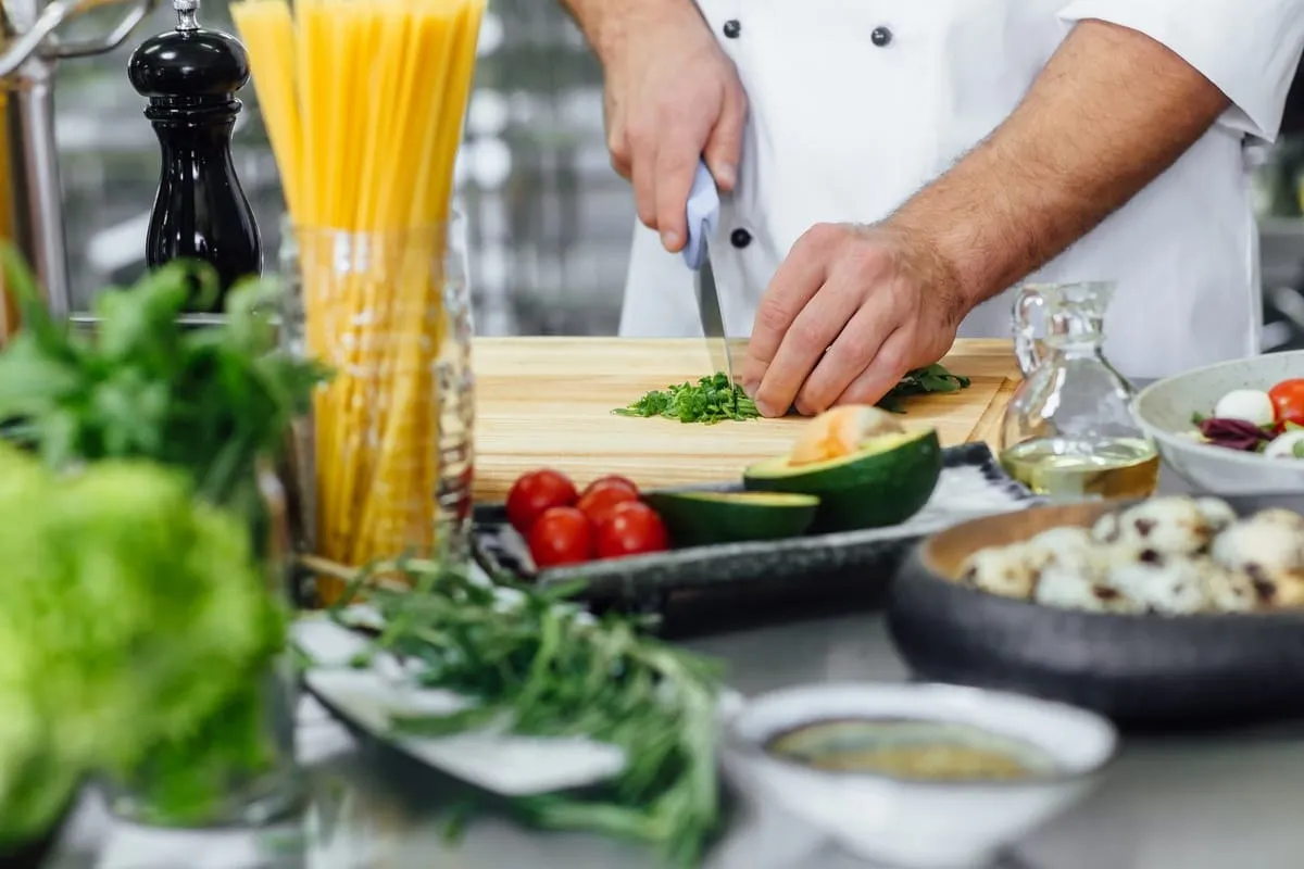 O persoană în haină albă taie verdeață pe un tocător, lângă paste, roșii, legume, recipiente cu ulei și o râșniță neagră.