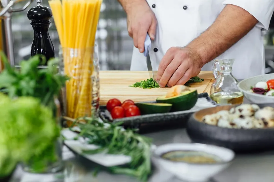 O persoană în haină albă taie verdeață pe un tocător, lângă paste, roșii, legume, recipiente cu ulei și o râșniță neagră.