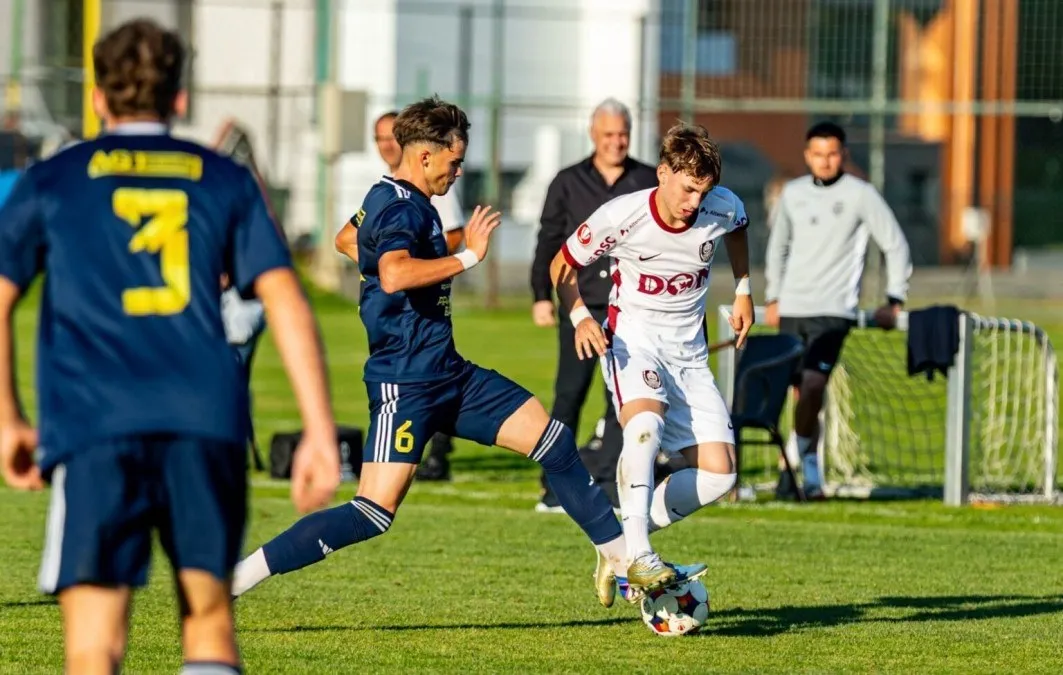Pe un teren cu iarbă, doi fotbaliști în echipamente alb și albastru urmăresc mingea, iar alți bărbați stau în prim-plan și în fundal.