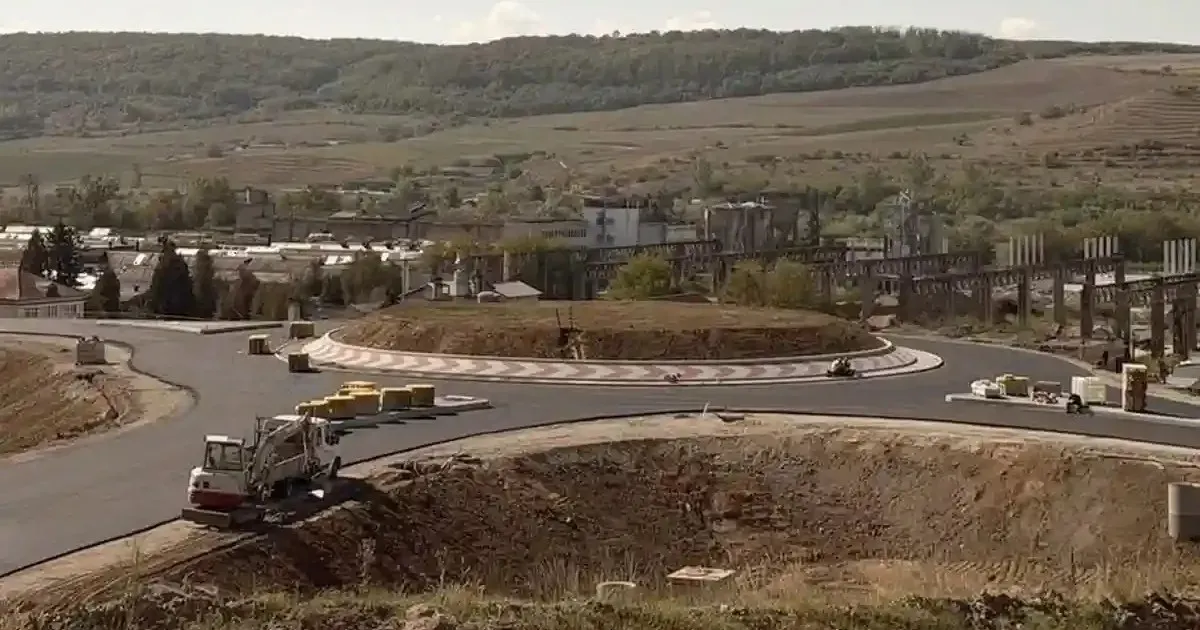 Un sens giratoriu în construcție este înconjurat de carosabil; în prim-plan se află un camion alb, iar în fundal se văd dealuri și clădiri.