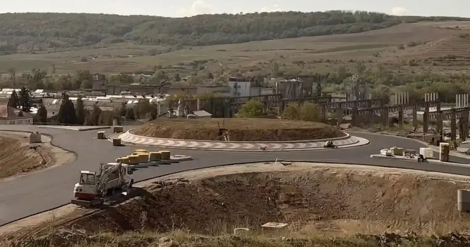 Un sens giratoriu în construcție este înconjurat de carosabil; în prim-plan se află un camion alb, iar în fundal se văd dealuri și clădiri.