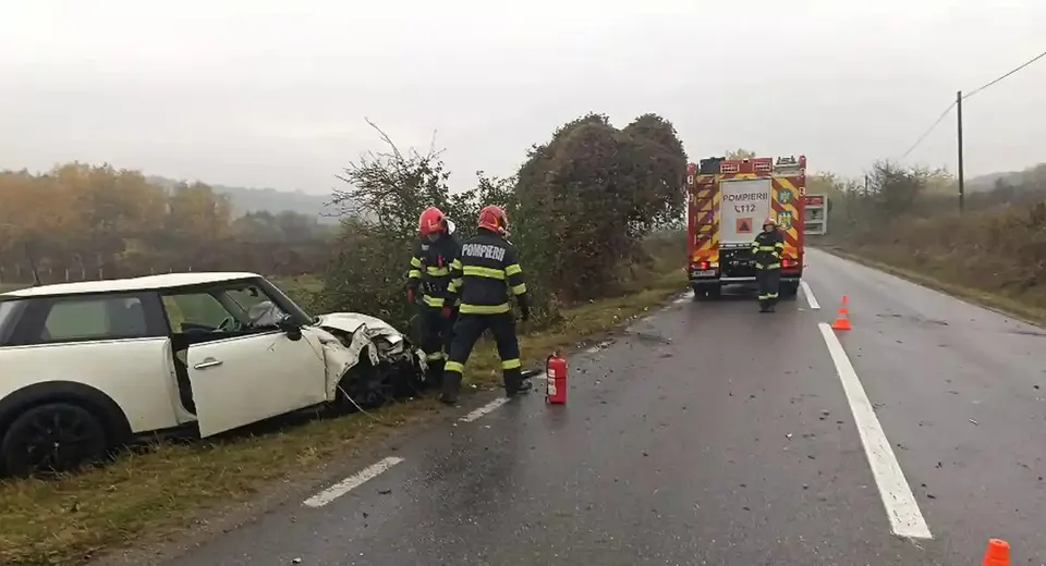 Un autoturism alb cu plafon negru și portiera deschisă are partea din față avariată; două persoane echipate stau lângă el, iar o autospecială este pe șosea.