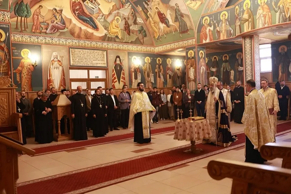 Mai mulți clerici în veșminte negre și aurii stau într-un interior pictat, lângă o masă acoperită, cu strane în prim-plan.