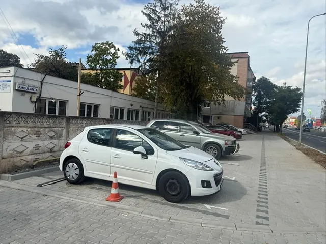 Un autoturism alb este parcat lângă un con portocaliu, cu alte mașini, o clădire joasă și o suprafață pavată în fundal.
