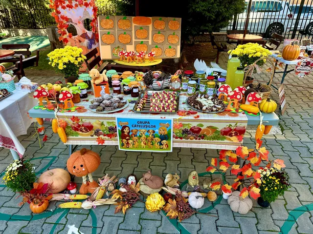 O masă acoperită cu pânză albă are dovleci decorați, borcane, tăvi cu prăjituri și flori; sub ea sunt așezați alți dovleci.