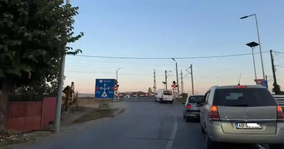 Pe o șosea, mai multe mașini se află în fața unor stâlpi și indicatoare, iar în stânga se văd copaci și un gard.
