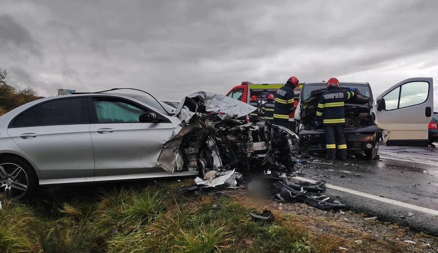Un autoturism argintiu are partea din față distrusă; fragmente sunt împrăștiate pe carosabil, iar două persoane în echipament de intervenție stau lângă vehicule.