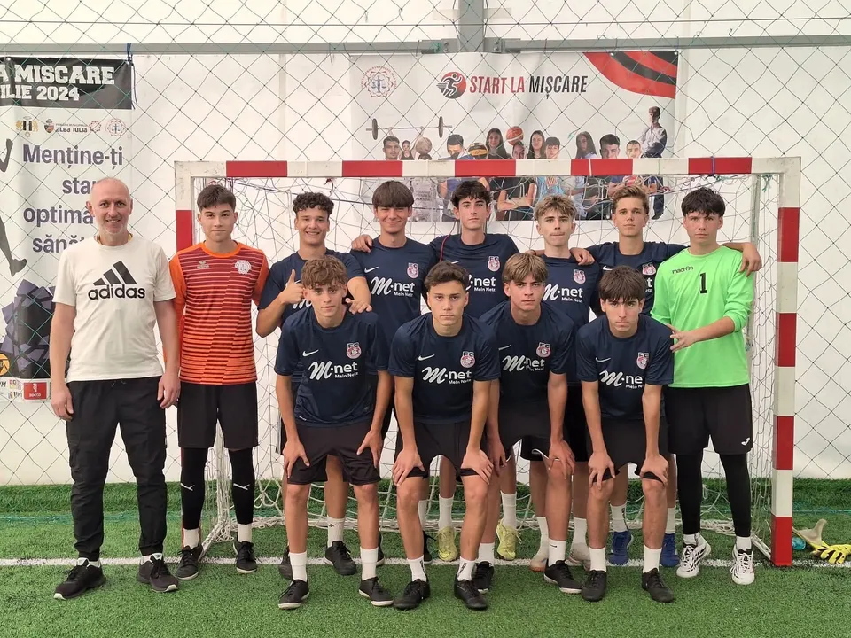Un grup de tineri în echipament sportiv și un adult în tricou alb pozează în fața unei porți de fotbal, pe teren artificial.