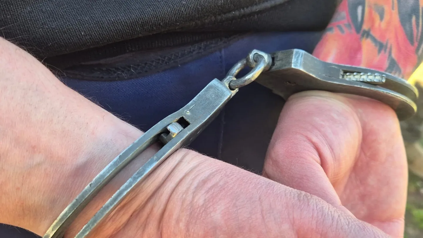 Două mâini apropiate sunt prinse cu cătușe metalice; în fundal se văd material textil închis la culoare și o suprafață cu model colorat.