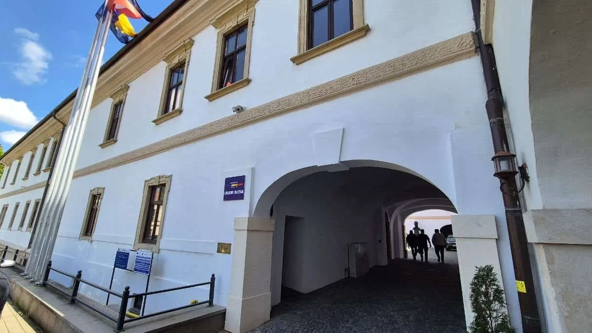 O clădire albă cu ferestre maro, steaguri, plăci albastre și o arcadă largă, prin care se văd câteva siluete.