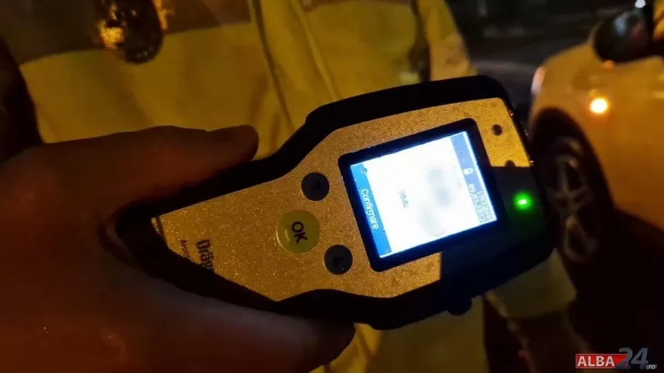 O mână ține în prim-plan un aparat cu ecran luminat și butoane; în spate se văd o persoană îmbrăcată în galben și un vehicul alb.