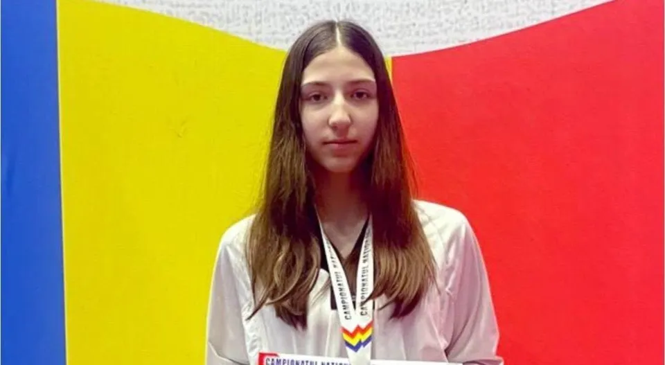 O tânără cu părul lung poartă o uniformă albă și o medalie la gât, în fața unor panouri colorate.