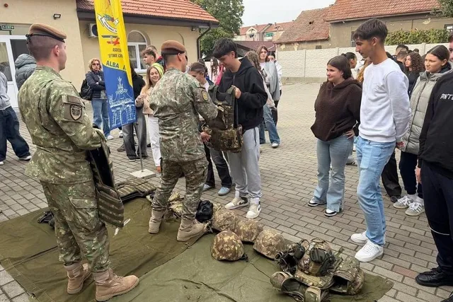 Doi bărbați în uniforme de camuflaj stau lângă mai mulți tineri, iar pe o prelată verde sunt așezate căști și alte echipamente.