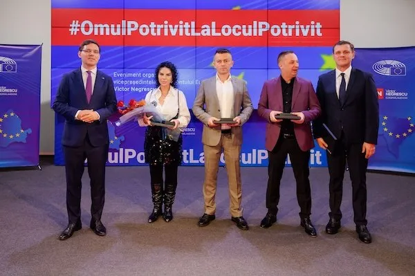 Cinci persoane stau în picioare în fața unui panou colorat; trei țin obiecte dreptunghiulare, iar patru poartă sacouri.