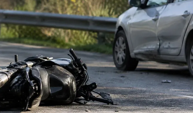 O motocicletă neagră este culcată pe carosabil, lângă un autoturism argintiu cu portiera laterală deformată; în spate se vede un parapet metalic.